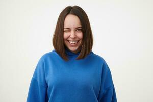 Cheerful young attractive short haired blue-eyed woman with bob haircut smiling gladly while winking at camera, dressed in blue hoodie while posing over white background photo