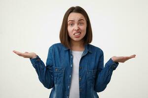 Indoor shot of bewildered young brown haired female with short haircut grimacing confusedly her face while standing over white background with raised palms photo