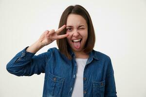 Indoos shot of joyful young brunette lady with natural makeup giving wink at camera and sticking out her tongue while raising hand with victory gesture, isolated over white background photo