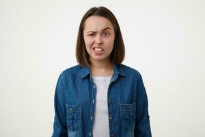 Displeased young short haired brunette female without makeup showing her teeth and raising confusedly eyebrow while looking at camera, isolated over white background photo