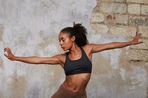 Indoor shot of young beautiful dark skinned curly female with belly button piercing rehearsing modern dance, posing over brick wall background with opened arms photo