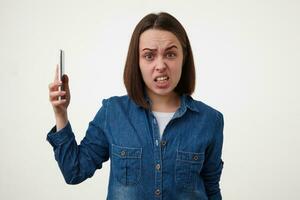 Aggressive young short haired lady with natural makeup grimacing her face while looking excitedly at camera, posing over white background with mobile phone in raised hand photo