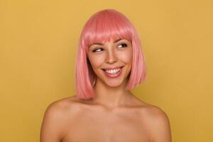 Portrait of young attractive pink haired female showing her perfect white teeth while smiling cheerfully, looking aside positively while standing over mustard background photo