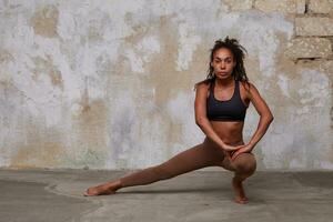 Image of young pretty athletic dark skinned lady with curly brown hair stratching her legs before dancing class, looking at camera with concentrated face, posing over loft interior photo