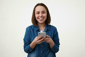 Indoor photo of young pretty cheerful dark haired lady smiling broadly while looking gladly at camera, holding mobile phone while posing over white background