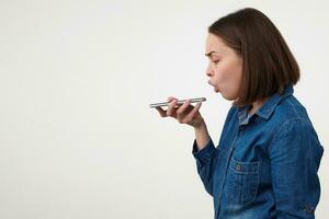 Studio photo of young short haired brunette lady dressed in blue jeans coat shouting at her smartphone while having tense conversation, posing over white background