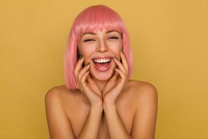 Joyful young attractive female with short pink hair wearing natural makeup while posing over mustard background, touching gently face with raised hands while laughing happily photo