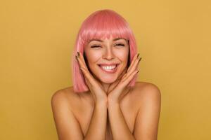 Happy young beautiful woman with pink trendy bob haircut keeping raised palms near her face and biting underlip while looking joyfully at camera, standing over mustard background photo