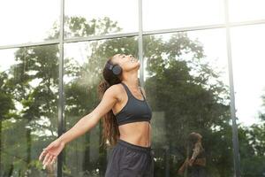 Positive smiling young fitness model with dark skin relaxing after hard workout with opened arms and closed eyes, dressed in black athletic wear while standing over urban background photo