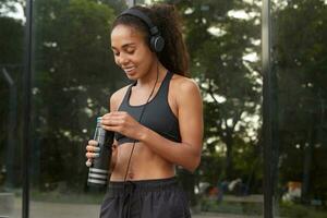 Plasant looking positive young sporty dark skinned female making break with training and listening to music with headphones. Healthy lifestyle, development and positive concept photo