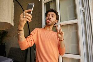 Funny indoor portrait of pretty bearded male holding phone in hand and leaning on window, making ridiculous faces and showing peace gesture, taking selfie with mobile phone photo