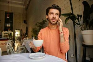 Young stylish bearded man posing over coffee house, talking on phone with cup of tea in hand and talking on phone, wearing peach color sweater photo