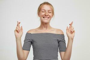 Good looking young lovely redhead woman dressed in striped t-shirt crossing her fingers while making wish with closed eyes and smiling cheerfully, standing against white background photo