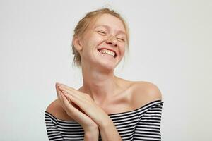 Pleased young attractive redhead female with natural makeup keeping raised palms together and smiling cheerfully with closed eyes, standing against white background photo