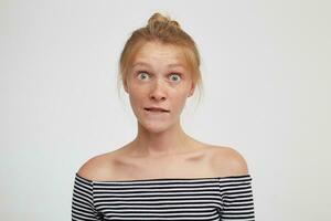 Amazed young attractive redhead lady with natural makeup rounding her green eyes and biting underlip while looking surprisedly at camera, isolated over white background photo