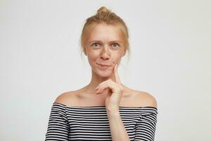 Portrait of young lovely green-eyed redhead female with casual hairstyle twising her mouth while looking thoughtfully at camera and keeping forefinger on her cheek, isolated over white background photo