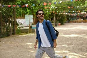 Outdoor shot of happy handsome young man with beard walking down green city park on sunny day, looking away with wide smile, wearing casual clothes photo