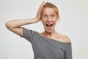 Indoor shot of overjoyed young lovely redhead female dressed in casual wear holding raised hand on her head and looking excitedly at camera with wide mouth opened, isolated over white background photo