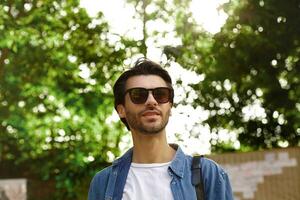 Outdoor close-up of charming young man with beard posing over green park on sunny day, wearing sunglasses and casual clothes, being in nice mood photo