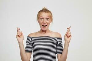 Agitated young attractive redhead female with bun hairstyle keeping her fingers crossed while looking excitedly at camera with wide eyes and mouth opened, posing over white background photo