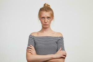 Upset young pretty redhead lady with bun hairstyle folding her lips while looking sadly at camera, crossing her hands while standing over white background photo