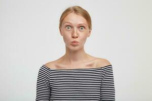 Suprirsed young pretty redhead female with natural makeup rounding her green eyes and folding lips while looking at camera, isolated over white background photo