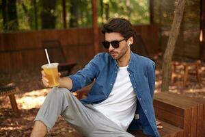 Young serious handsome man in casual wear having lunch break, holding glass of ice tea while sitting in public garden, listening to music with earphones photo