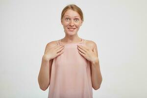 Cheerful young lovely redhead lady with natural makeup looking excitedly at camera with wide eyes opened and holding palms on her chest while posing over white background photo