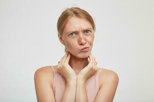 Puzzled young attractive redhead lady frowning her eyebrows and twisting mouth while looking confusedly at camera, keeping raised hands on neck while posing over white background photo