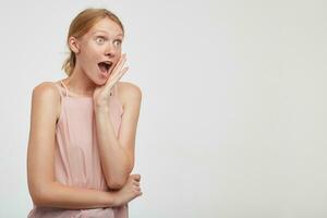 Amazed young pretty redhead woman holding raised palm on her cheek and looking surprisedly aside with wide eyes and mouth opened, wearing casual clothes over white background photo