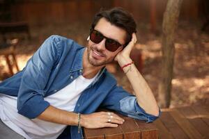 Outdoor shot of attractive young man with beard leaning on his head with palm, posing over city garden with sincere smile, wearing casual clothes photo