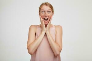 Amazed young pretty redhead female dressed in pink shirt holding raised palms on her cheek and looking surprisedly at camera with wide eyes opened, isolated over white background photo