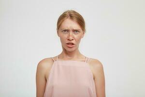 Confused young green-eyed redhead female grimacing her face while looking perplexedly at camera and frowning her eyebrows, isolated over white background photo