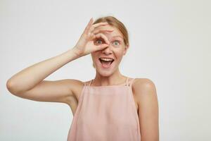 Funny shot of young attractive redhead female with casual hairstyle raising ok gesture to her eye and looking joyfully at camera with opened mouth, isolated over white background photo