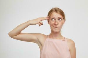 Puzzled young attractive redhead lady in pink top looking confusedly aside and twising her mouth while keeping gun gesture on her temple, isolated over white background photo