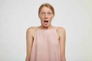 Astonieshed young pretty redhead female rounding her green eyes while looking amazedly at camera with wide mouth opened, isolated over white background photo