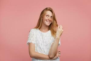 Cheerful young attractive redhead female pulling her hair with raised hand and looking dreamily upwards with wide happy smile, standing over pink background photo