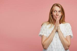 Indoor photo of young attractive redhead lady dressed in white festive t-shirt raising folded palms to her mouth while looking thoughtfully upwards, isolated over pink background