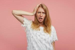 Puzzled young pretty redhead female lady with grey-green eyes keeping raised palm on her head and frowning eyebrows while looking confusedly at camera, isolated over pink background photo