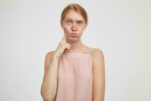 Studio shot of funny young redhead woman with natural makeup pursing her lips and making faces while posing over white background with raised hand in casual wear photo