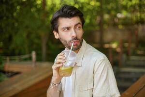 Outdoor portrait of charming young bearded male with dark hair looking aside and drinking lemonae with straw, posing over green city garden photo