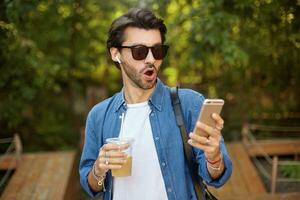 Surprised attractive young bearded man in sunglasses standing over green park with mobile phone in hand, receiving unexpected news, wearing casual clothes photo