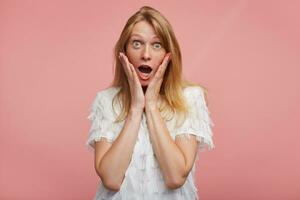 Shocked young pretty redhead woman holding her face with raised hands and looking amazedly at camera with wide eyes and mouth opened, isolated over pink background photo