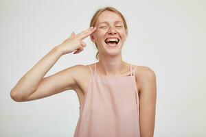 Good looking young cheerful redhead female folding gun with her fingers and keeping it near temple, laughing happily with closed eyes while posing over white background photo