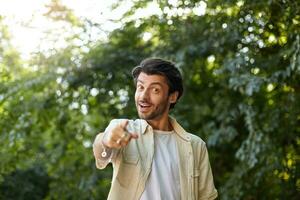 al aire libre Disparo de encantador joven barbado masculino con de moda Corte de pelo posando terminado verde arboles en soleado día, punzante a cámara con elevado dedo índice, siendo positivo y sonriente foto