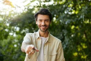 Portrait of cheerful young dark haired man looking to camera with sincere smile and raised forefinger, wearing casual clothes, posing over green park photo