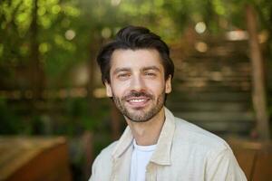 Close-up of beautiful bearded male with dark hair posing over green ciy park on sunny day, looking to camera with charming and sincere smile photo