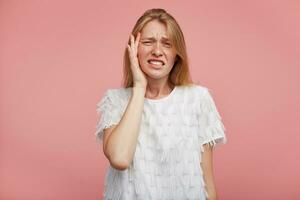 Portrait of sad young pretty woman with foxy hair showing her teeth while looking unhappily at camera and holding raised hand on her face, standing over pink background photo