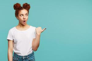 Surprised young brown haired curly lady with bun hairstyle rounding her mouth while looking amazedly aside and keeping her hand raised, isolated over blue background photo