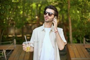 Young attractive bearded man in beige shirt and sunglasses making call with his mobile phone, walking around green city prk with lemonade in hand photo